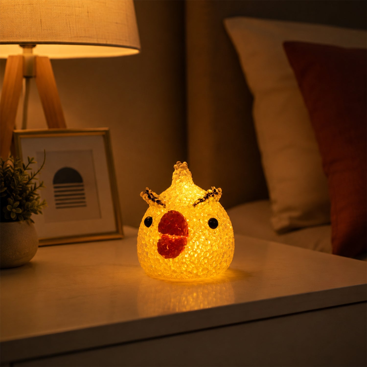 A yellow chick-shaped crystal texture LED lamp glowing on a white nightstand next to a lamp and framed picture.