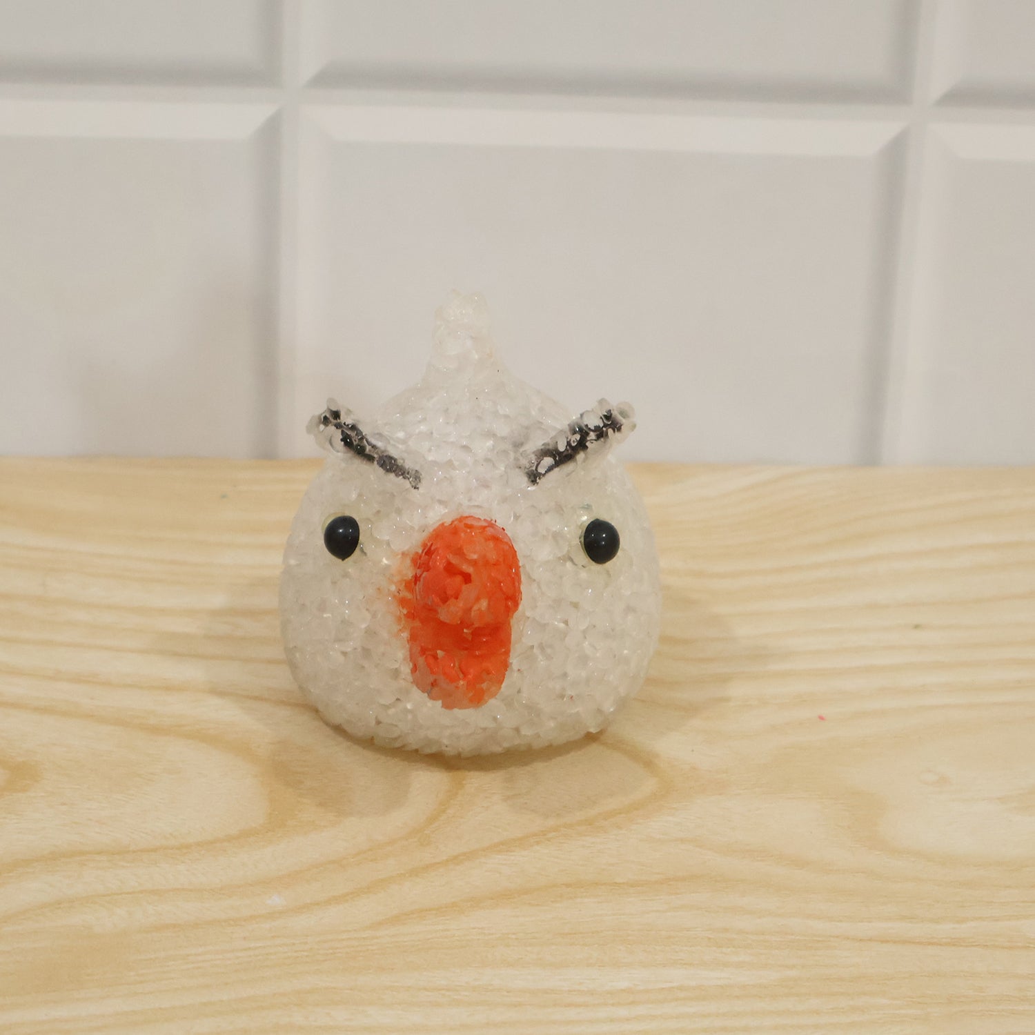 A white, unlit chick-shaped lamp with an orange beak and black eyes sits on a light wooden surface against a white tiled