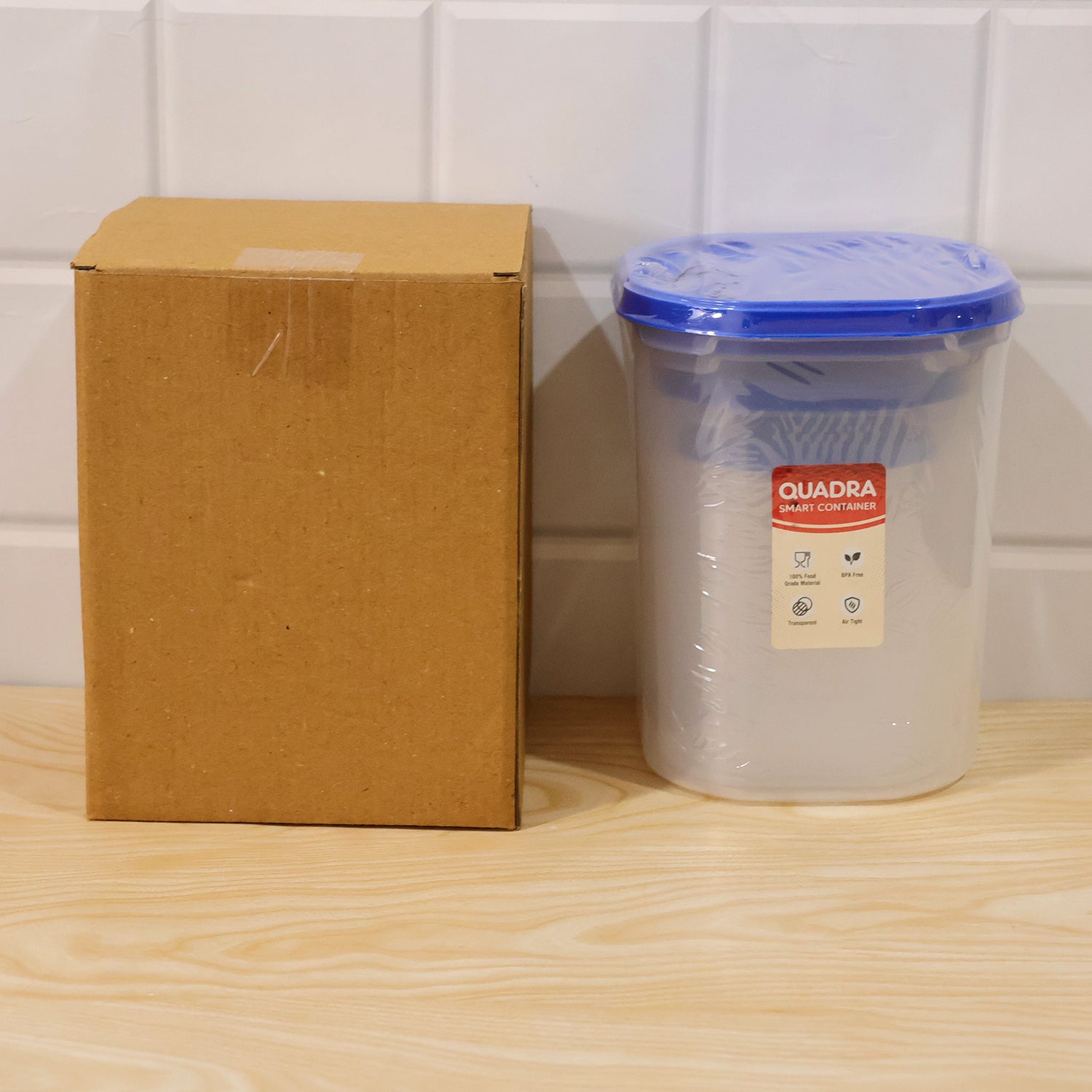 A brown cardboard box next to a single clear plastic container with a blue lid, wrapped in plastic, on a wooden surface.