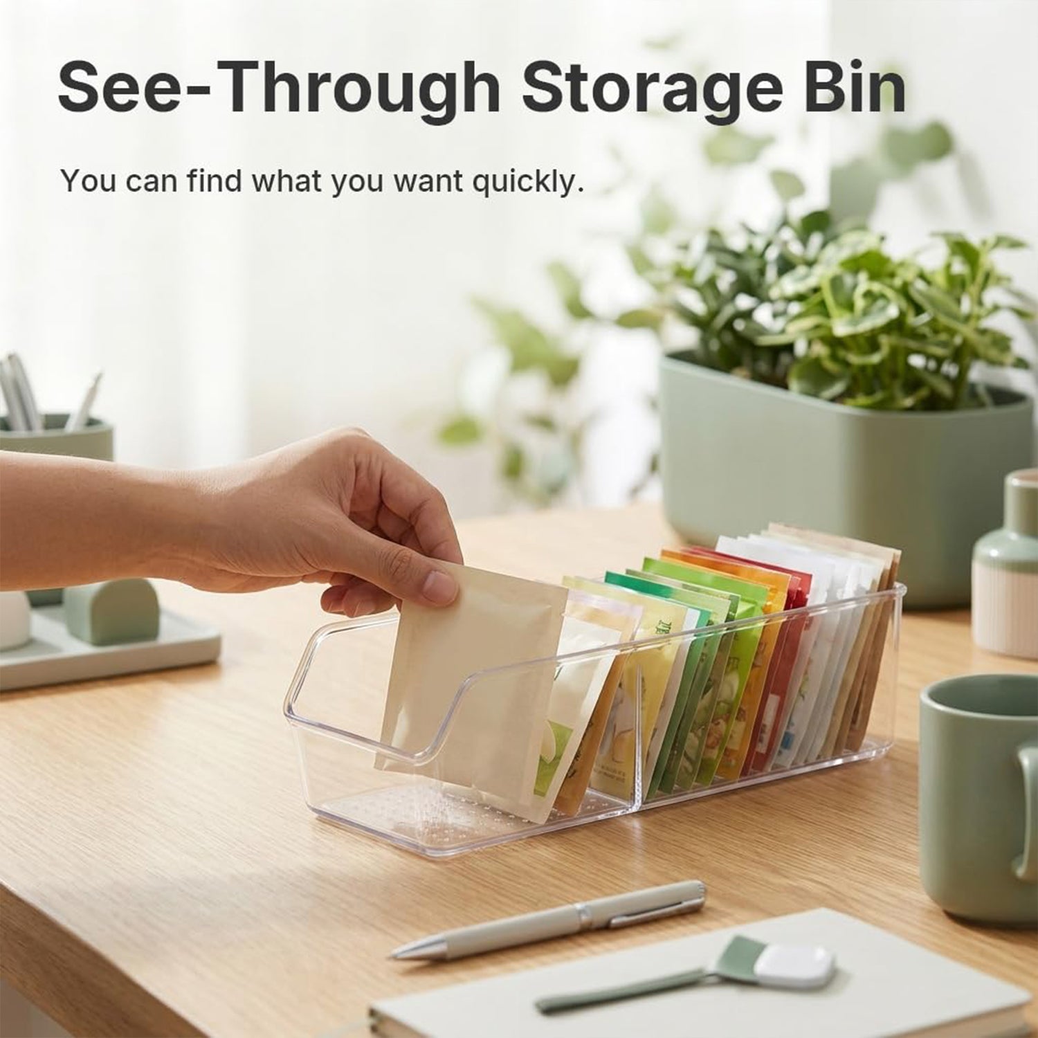 A hand retrieves a tea bag from a clear plastic organizer filled with colorful packets on a wooden desk with plants.