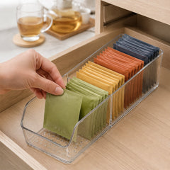 A hand places a green tea bag into a clear plastic organizer filled with colorful tea bags inside a wooden drawer.