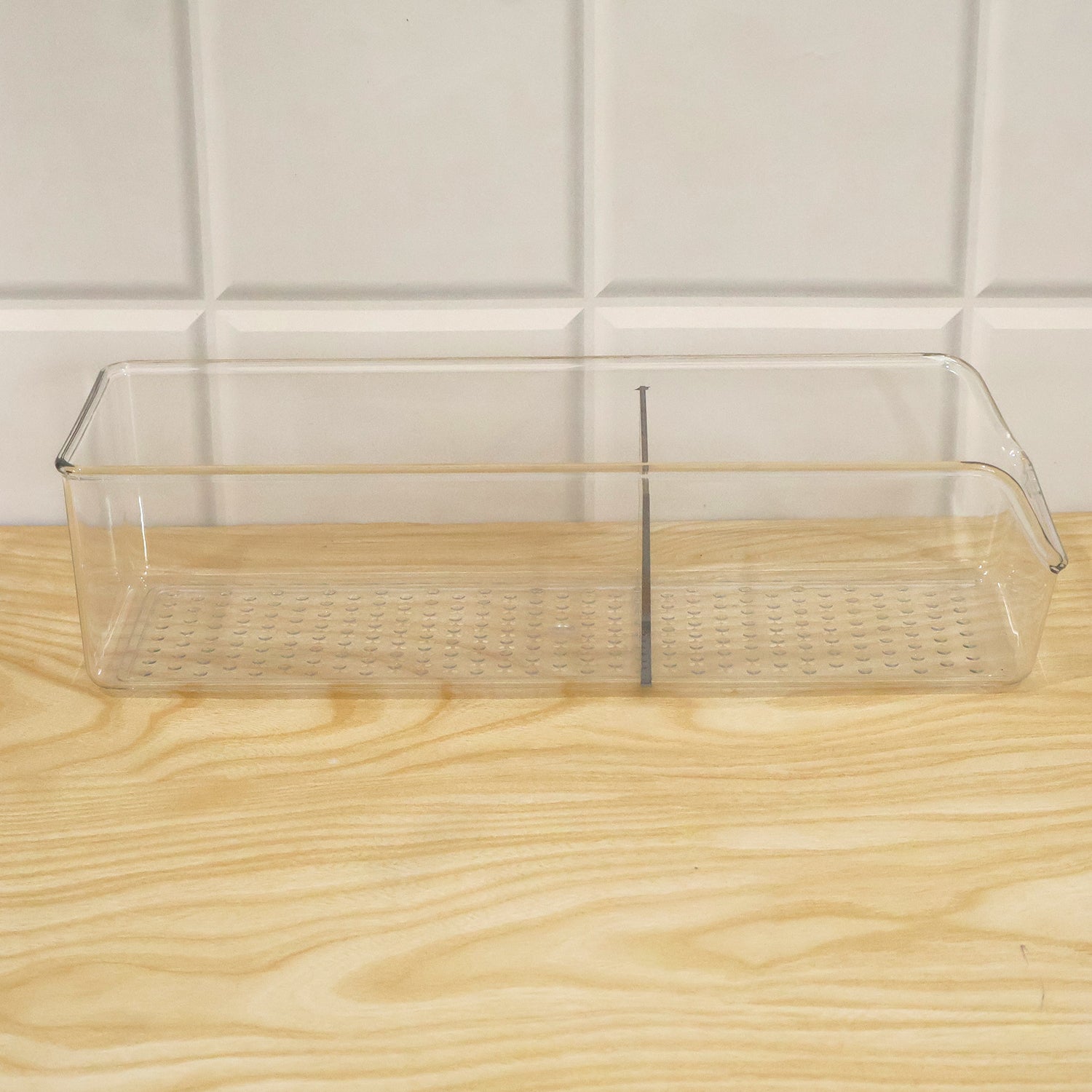 Empty clear plastic organizer with two sections and a textured bottom, sitting on a light wooden surface against white tiled
