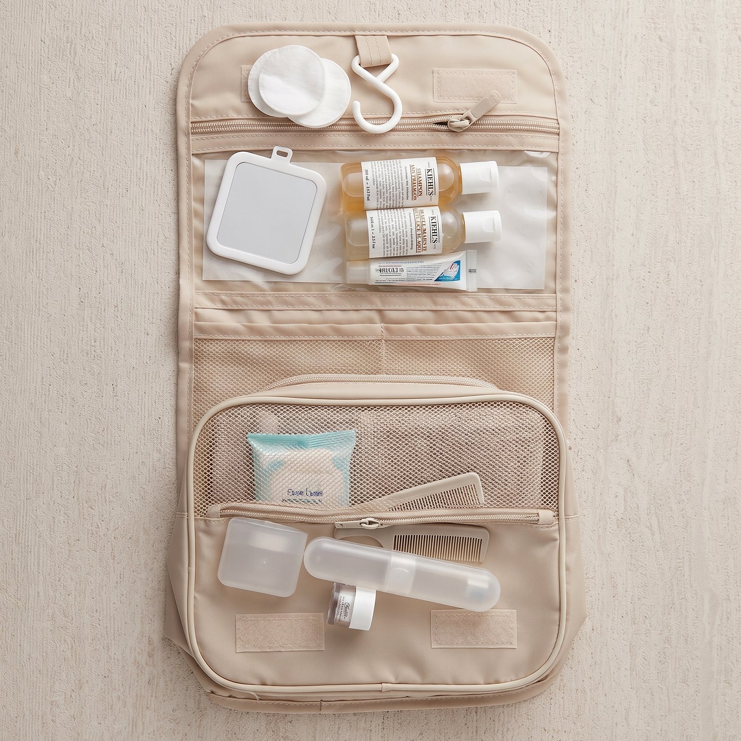 An open beige toiletry bag containing a mirror, small bottles, cotton pads, and a comb on a wooden surface.