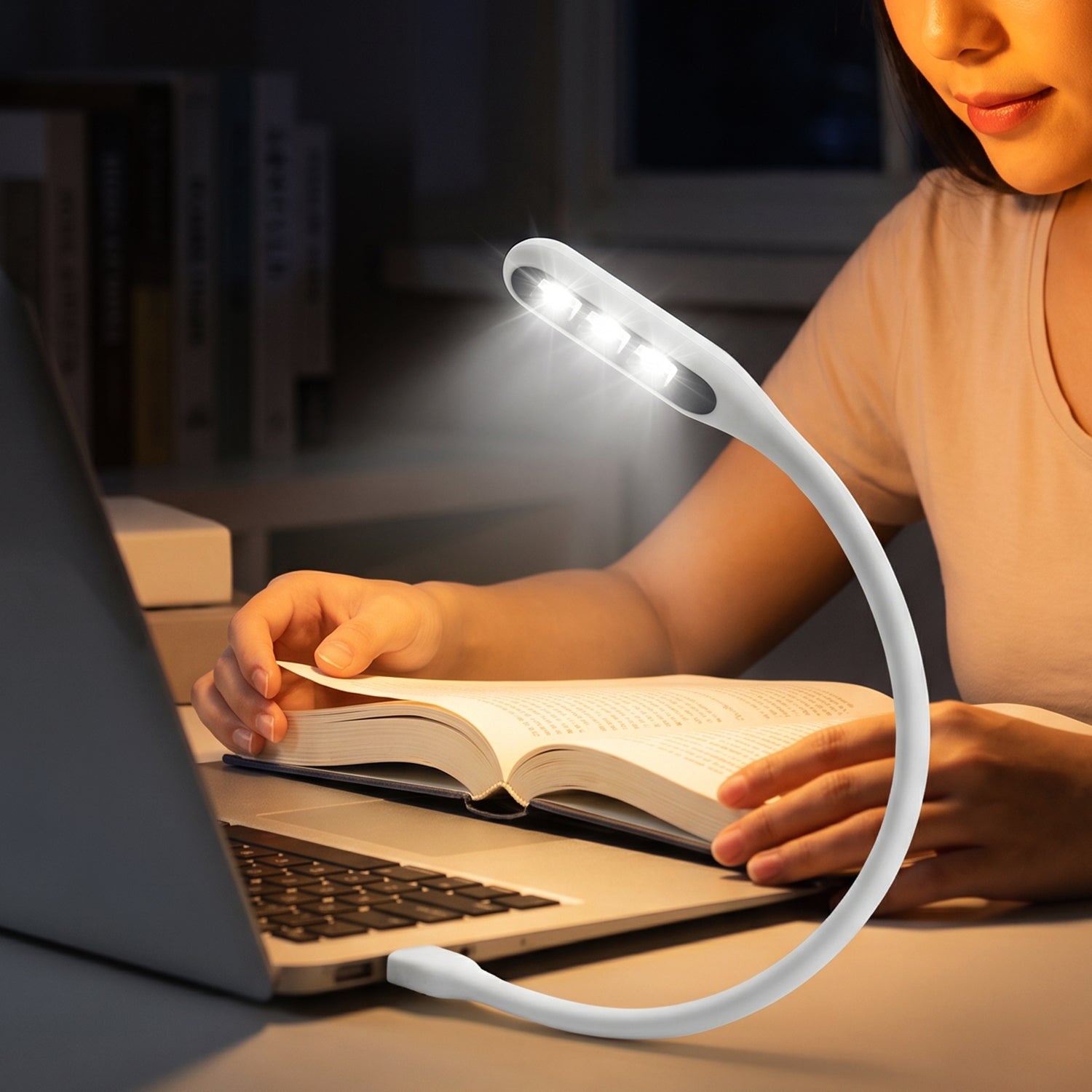 A person reading a book at night with a white flexible USB LED light plugged into a laptop, providing illumination.