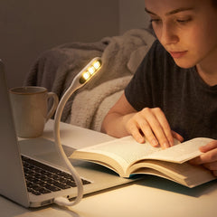 A person reads a book in a dimly lit room, illuminated by a white flexible USB LED light plugged into a laptop.