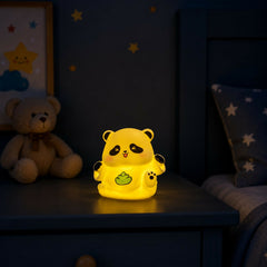 The panda night light glows yellow in a dimly lit room, illuminating a small area on a dark blue bedside table next to a