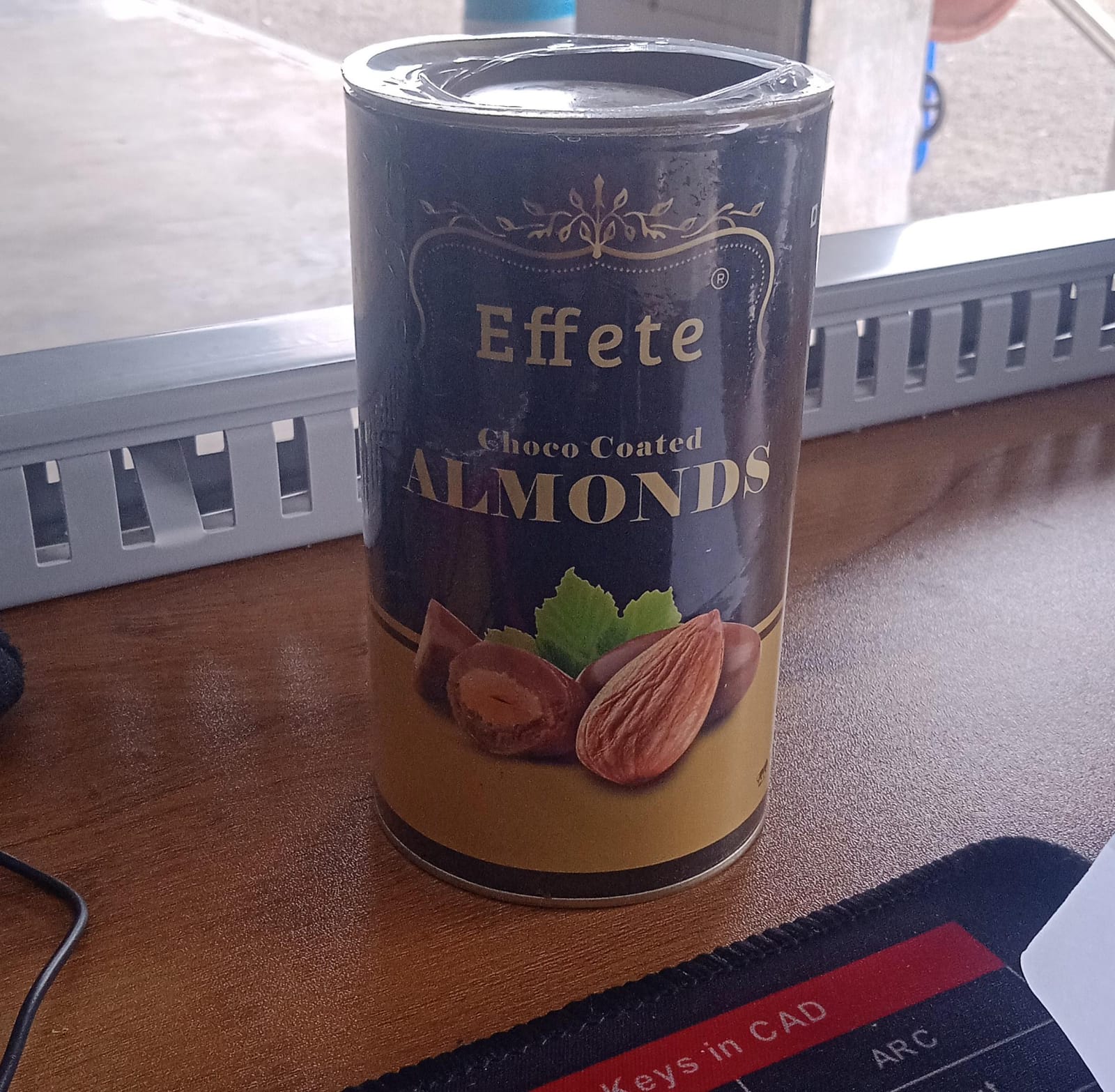 Effete Choco Coated Almonds tin on a wooden desk next to a black mousepad with white text.