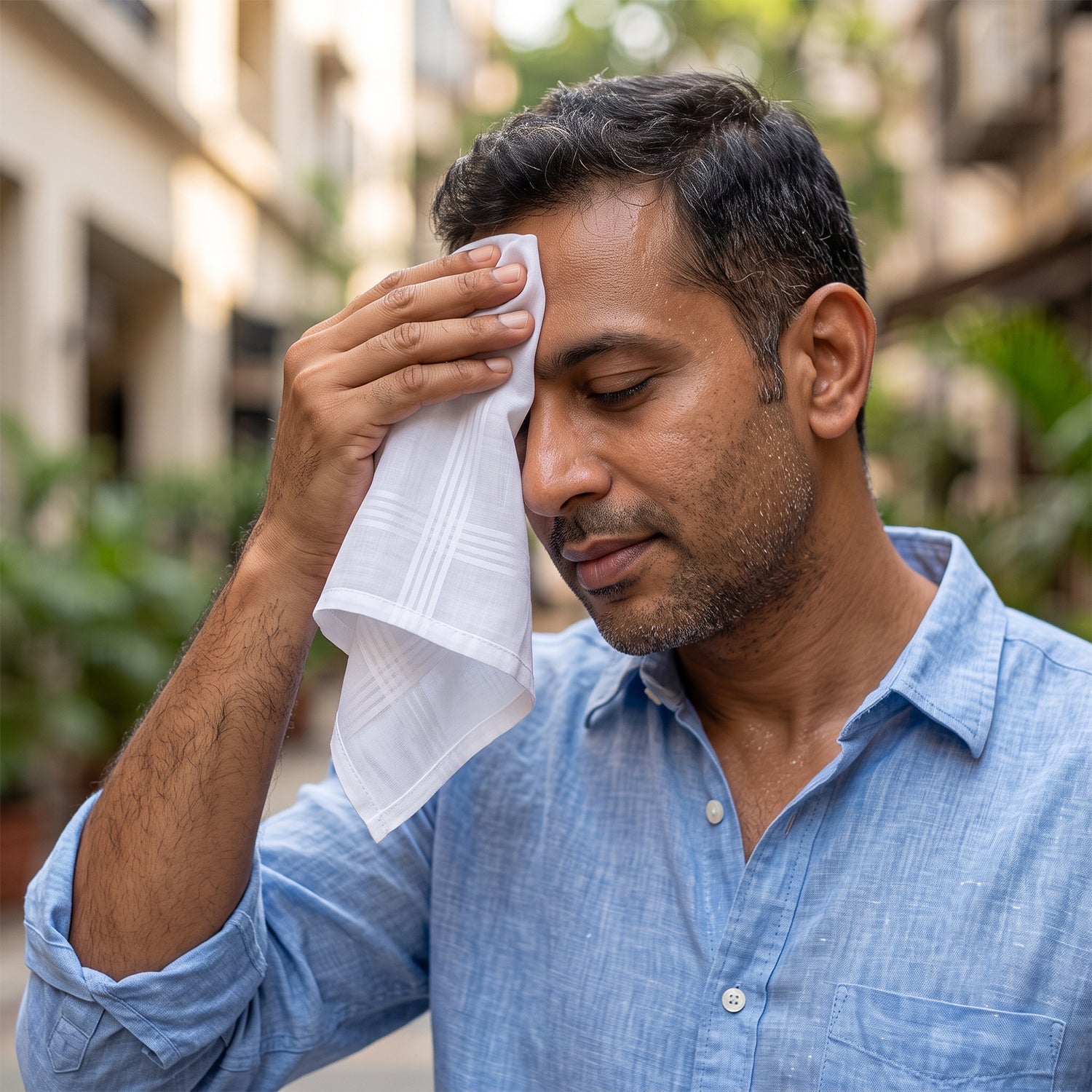 A man in a blue shirt wipes sweat from his forehead with a white cotton handkerchief on a sunny street.