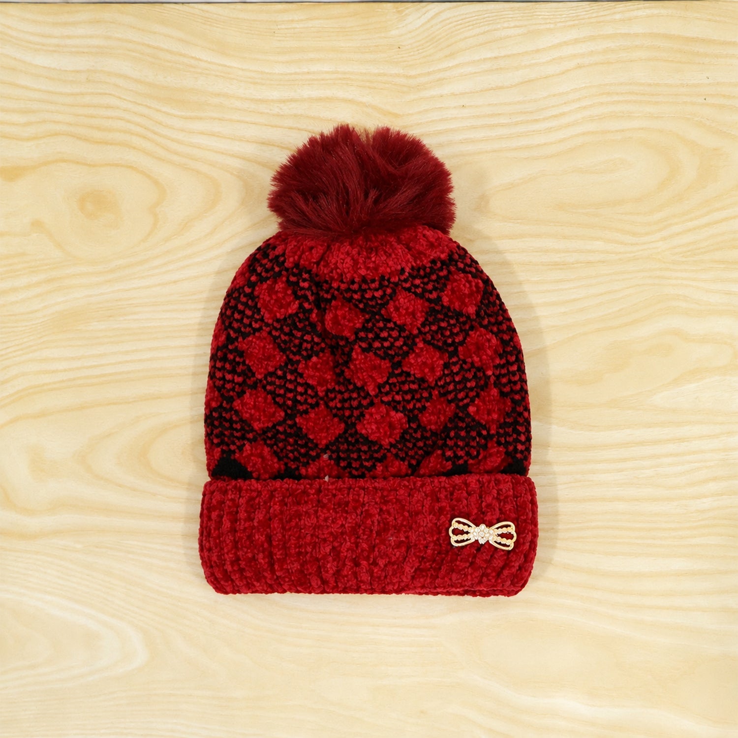 A red and black patterned beanie with a fluffy pom pom and a small bow detail is displayed on a light wooden surface.