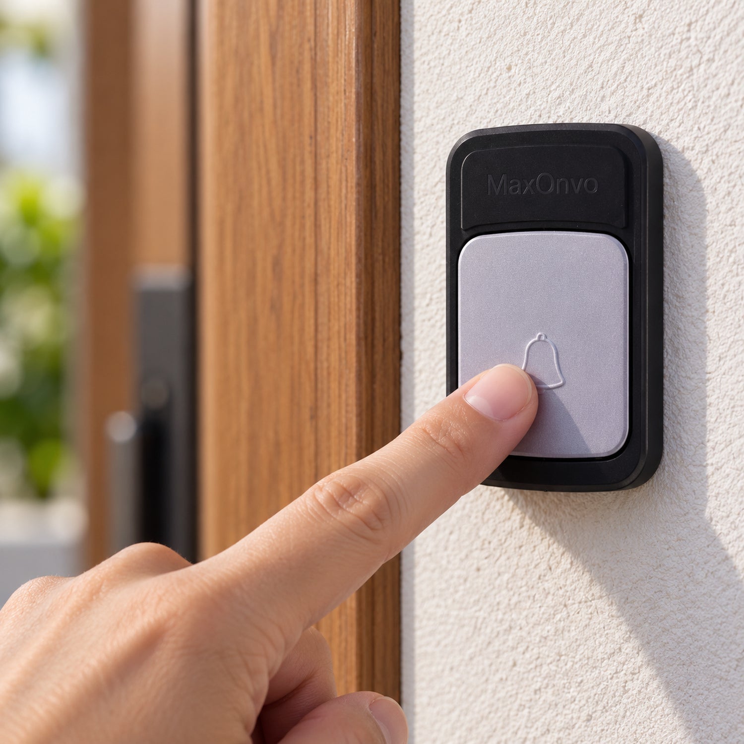 A person's finger presses the light grey button of a black wireless doorbell transmitter on a white wall next to a â€“ view 2