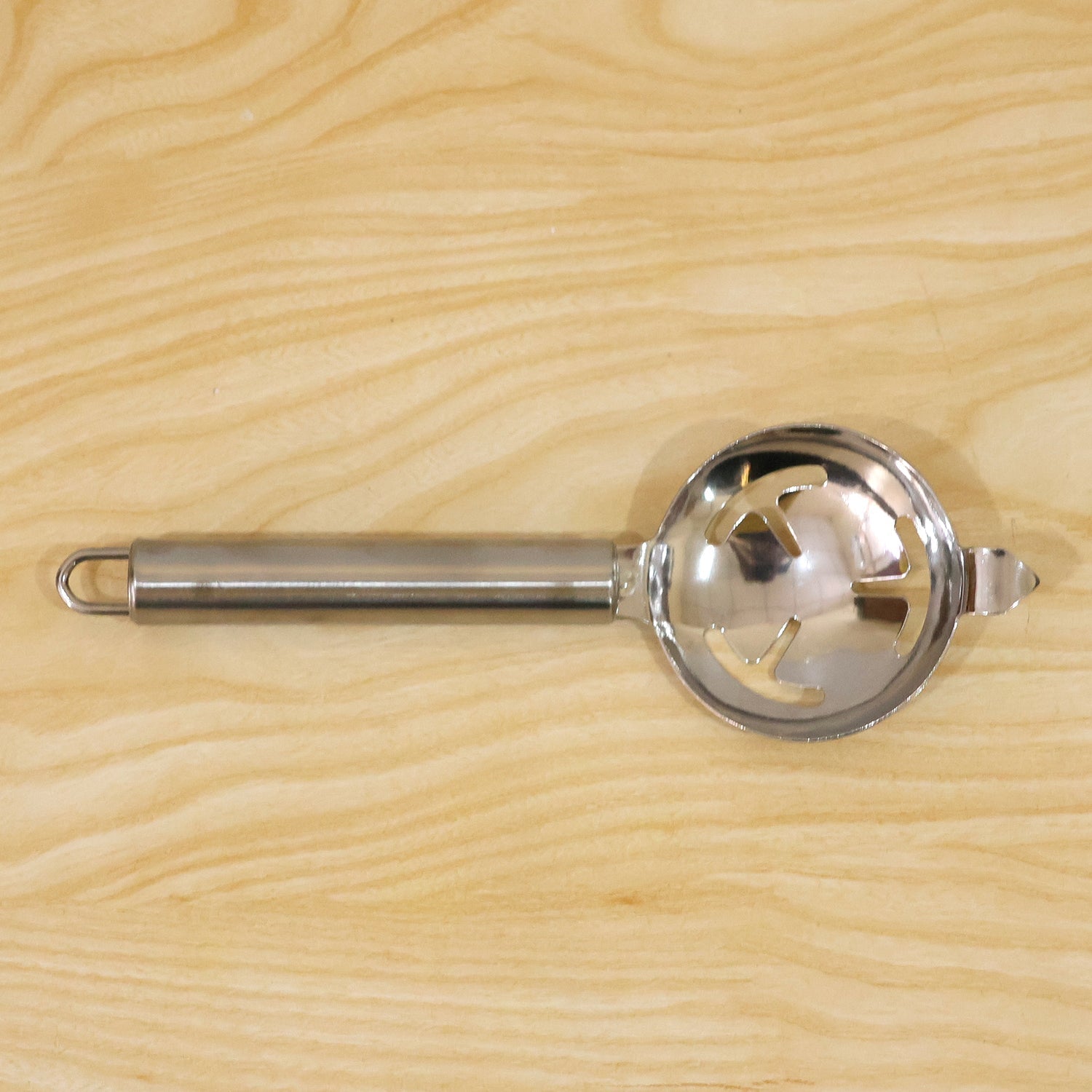 The stainless steel egg separator with its slotted bowl and long handle is centered on a light wooden background.