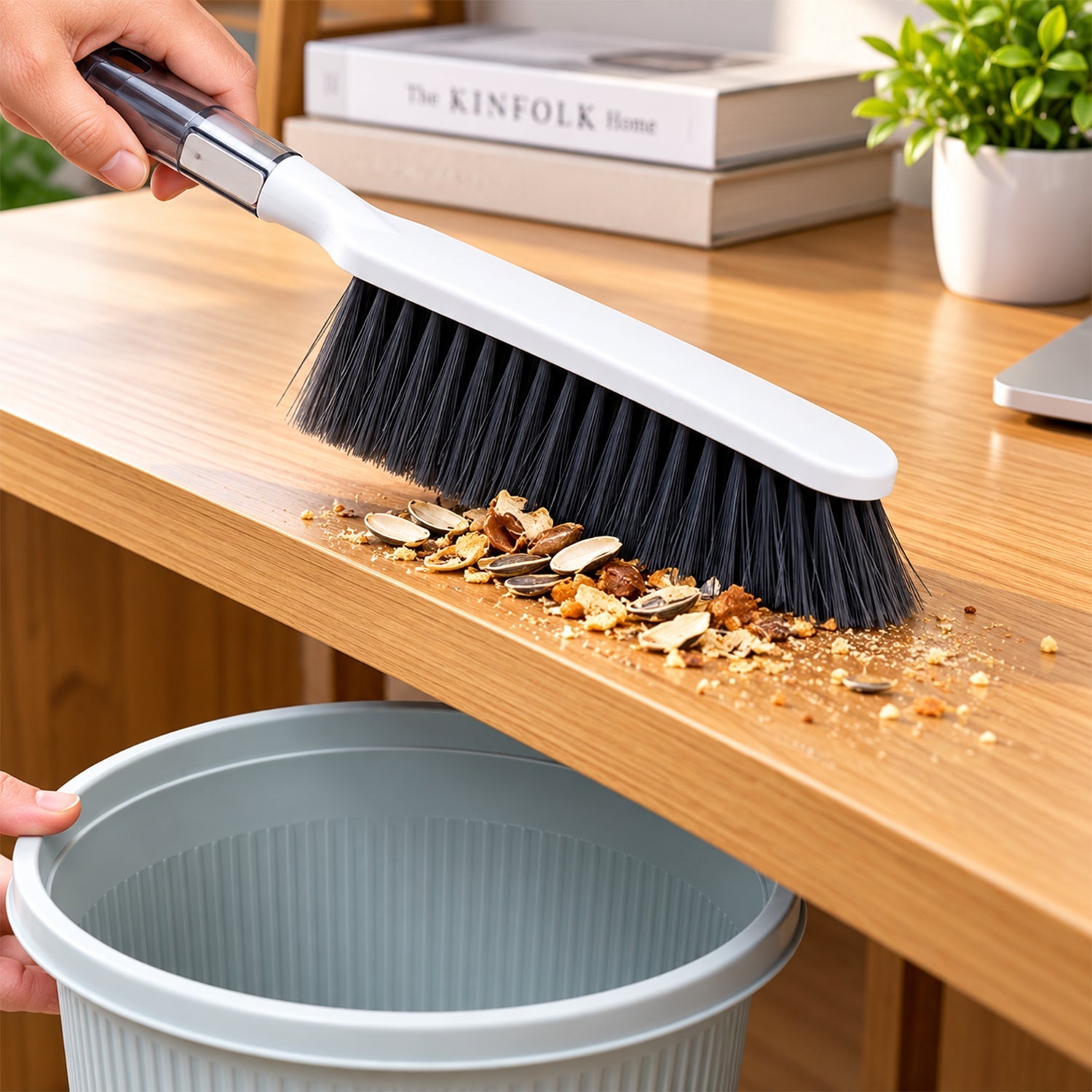 A hand holds the brush, sweeping a pile of seeds and crumbs from a wooden table toward a gray trash can.