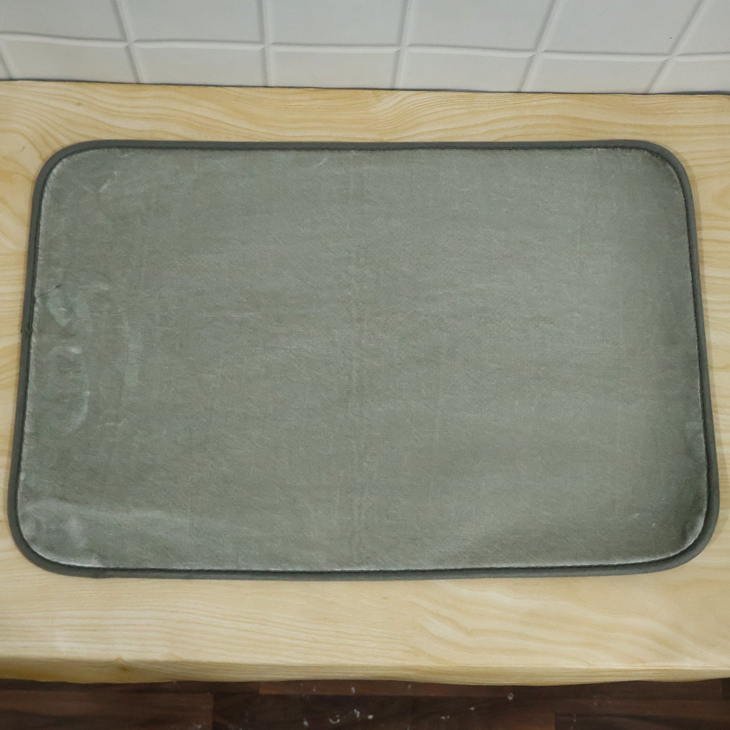 A gray rectangular floor mat resting on a light wooden surface with a white tiled wall behind it.