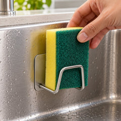 A hand places a yellow and green sponge into a single brushed metal holder attached inside a stainless steel sink basin.