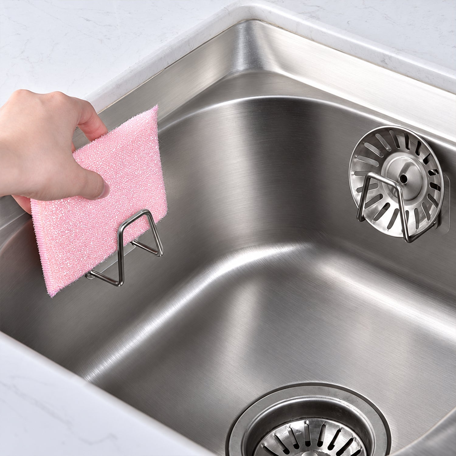 A hand places a pink scrubbing pad into a single stainless steel holder inside a sink, beside a sink strainer.