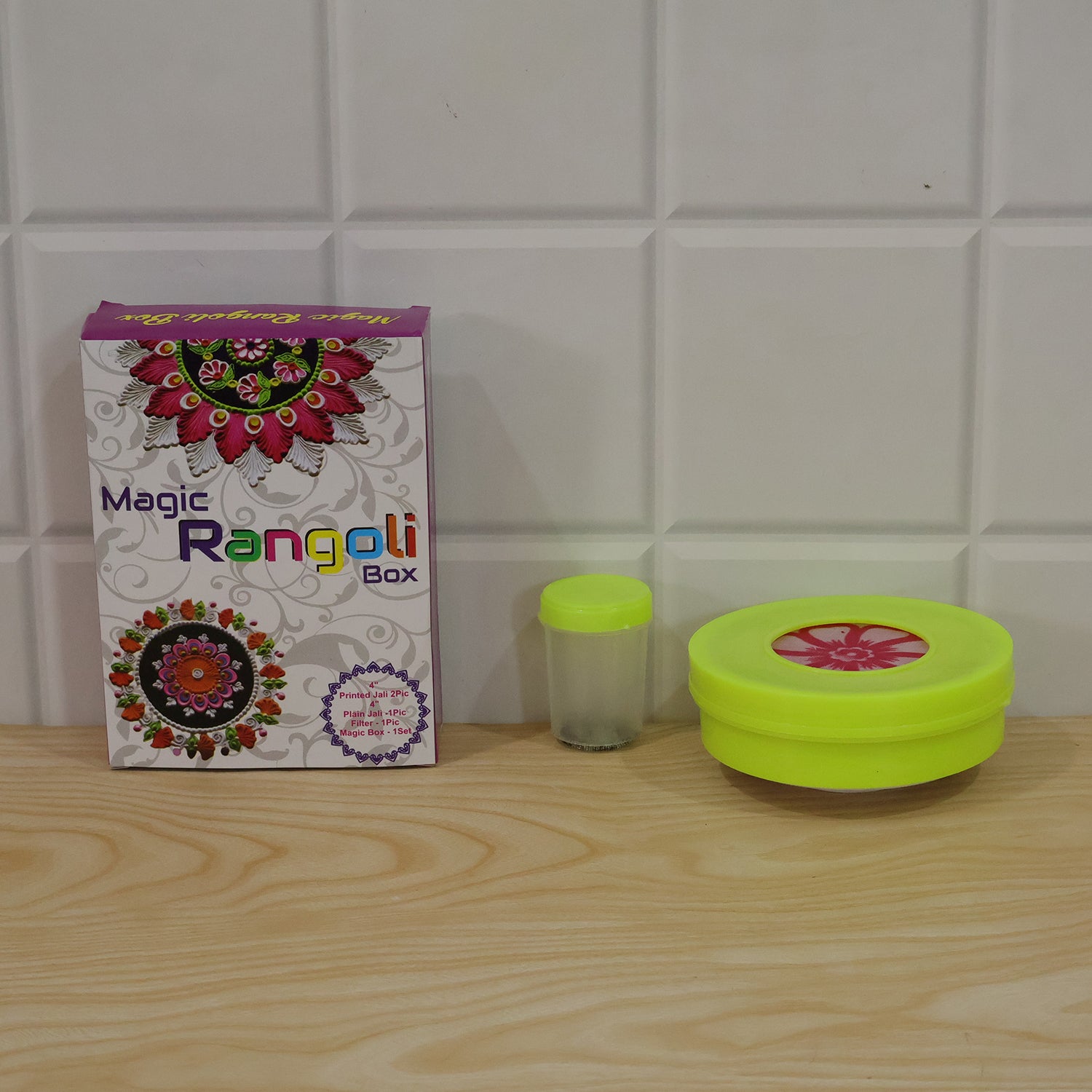 The Magic Rangoli Box, a small clear container, and a lime green powder dispenser are arranged on a wooden surface.