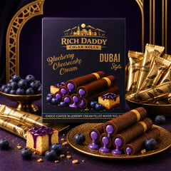 A decorative scene with a Rich Daddy box, gold-wrapped wafer rolls, fresh blueberries in bowls, and cheesecake pieces on a